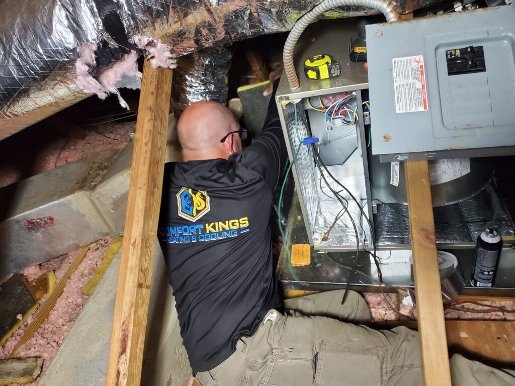 A technician wearing a "Comfort Kings Heating & Cooling" shirt works on HVAC equipment in an attic space surrounded by insulation and electrical components.