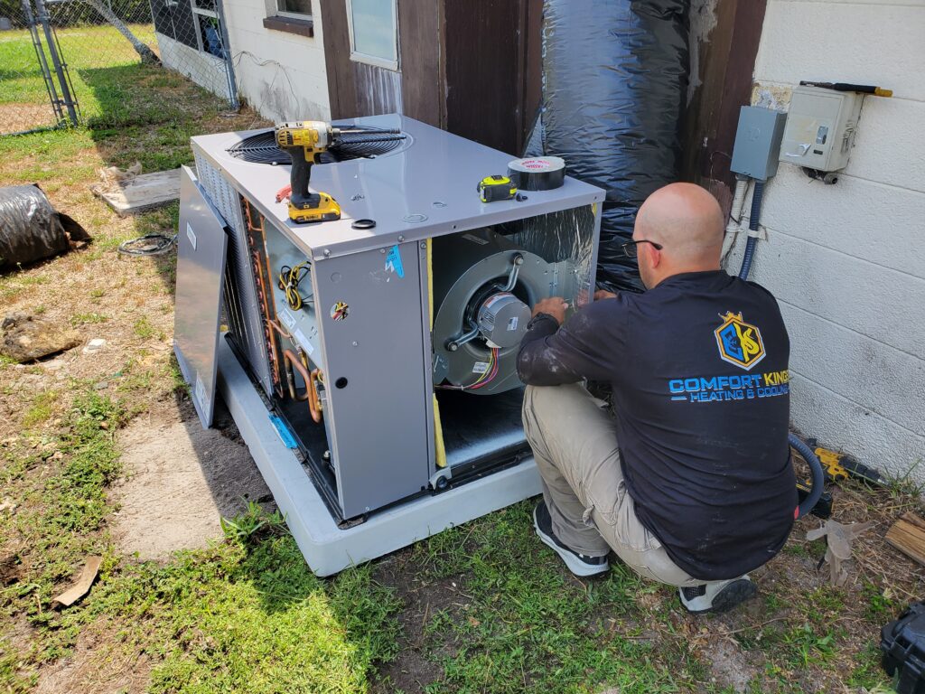 A technician in a "Comfort King Heating & Cooling" shirt services an outdoor HVAC unit next to a building, with tools placed on top of the unit.