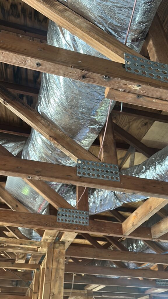 Exposed wooden ceiling beams with metal brackets and large silver HVAC ductwork running through a building under construction or renovation.