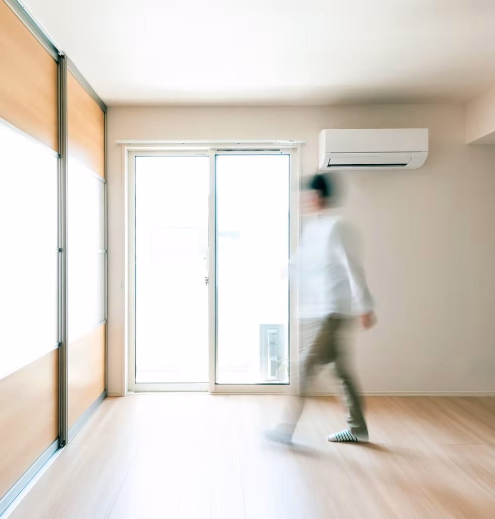 A person walks through a bright, empty room with wooden floors, large windows, sliding glass door, and a wall-mounted air conditioner.