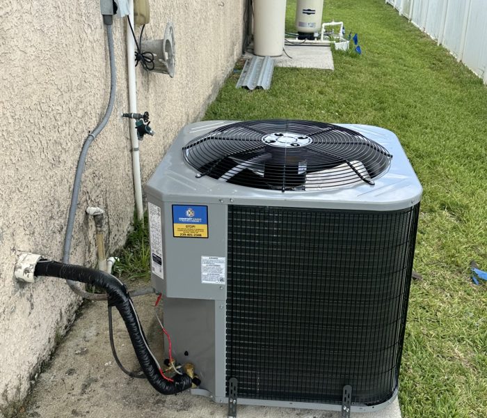 Outdoor air conditioning unit installed on a concrete pad beside a tan stucco house, with electrical wiring and pipes connected, and grass surrounding the area.